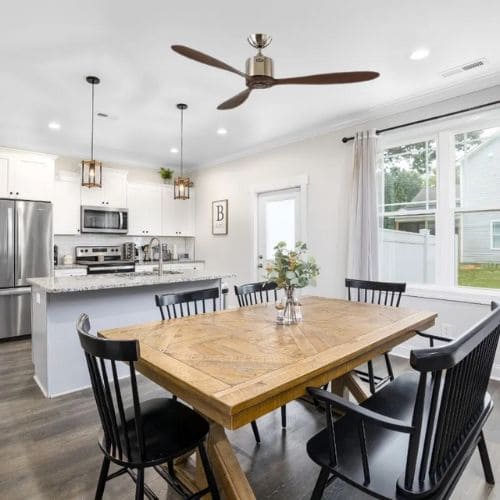 Ventilateur de plafond au-dessus d'une salle à manger moderne, grande table en bois et chaises noires, cuisine lumineuse en fond. sens de rotation ventilateur de plafond en hiver indiquant redistribution de chaleur.