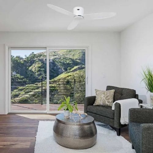 Ventilateur de plafond blanc dans un salon lumineux avec vue extérieure, canapé gris, plantes et table basse ronde en métal.