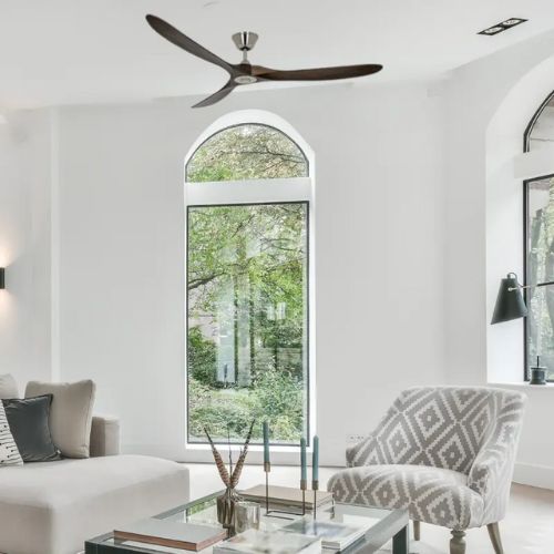 Ventilateur de plafond moderne installé au-dessus d’un salon lumineux avec canapé et fauteuil près d’une grande fenêtre.